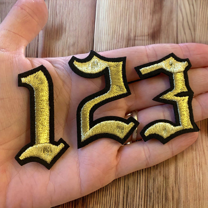 Metallic gold and black old english font iron on number patches showing the numbers 1, 2, 3 in a hand.