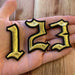 Metallic gold and black old english font iron on number patches showing the numbers 1, 2, 3 in a hand.