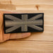 A hand holding a grey metallic thread embroidered Union Jack GB Flag sew-on patch with a hook and loop fastener backing on a wooden background