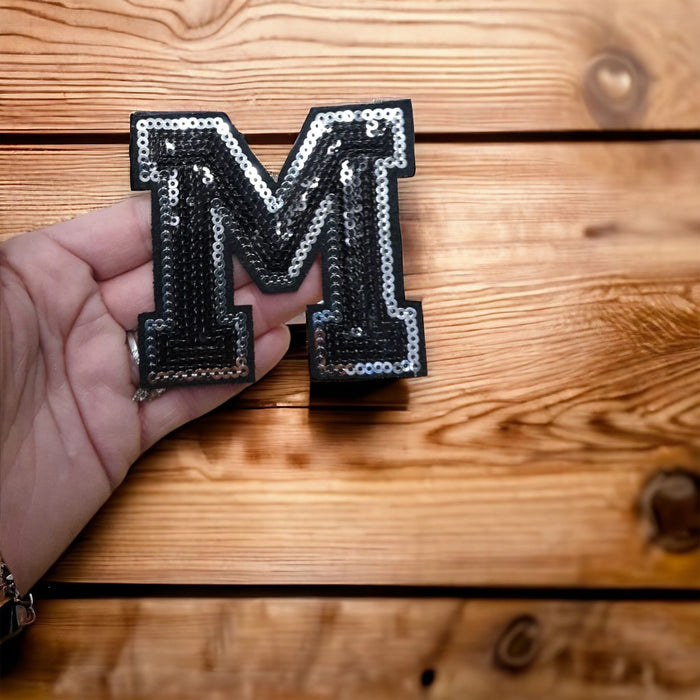 Black and silver sequin iron on patch letters 7.5cm height in a hand on wooden background